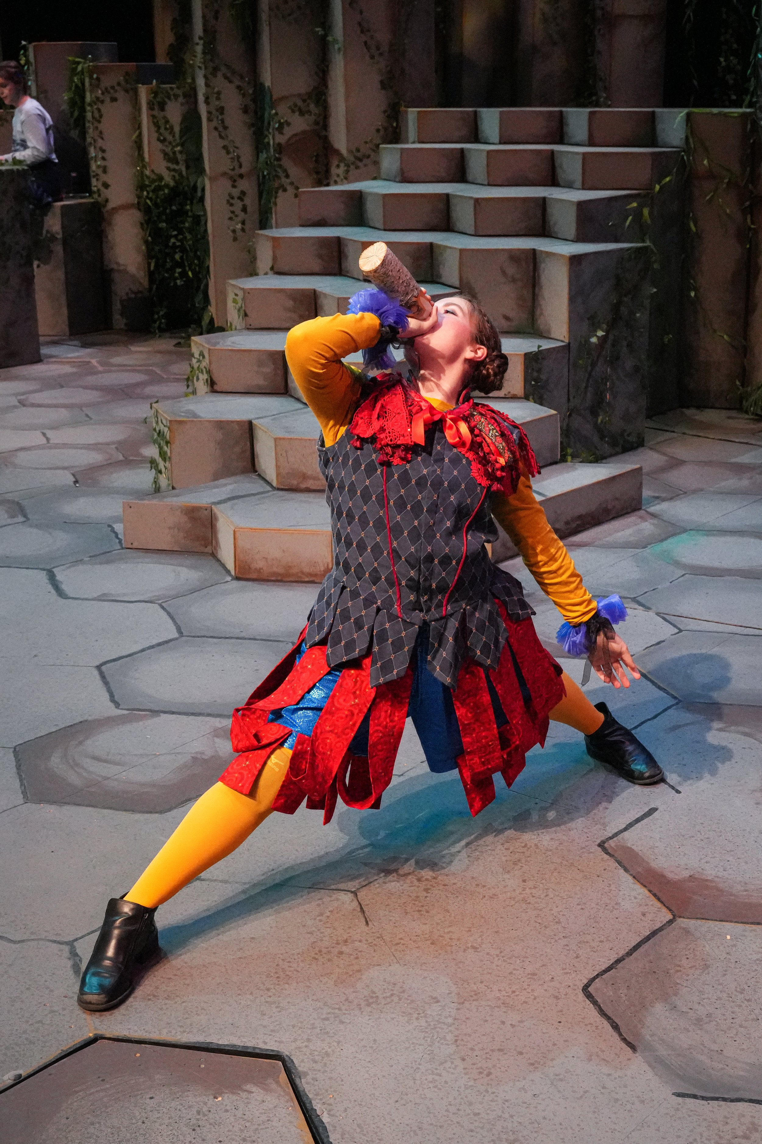 A woman in colorful costume, including a red ruffled collar, a red, black, and blue skirt, and yellow tights, is standing in a dramatic pose as she drinks from a prop bottle on a stage with stone steps and greenery.