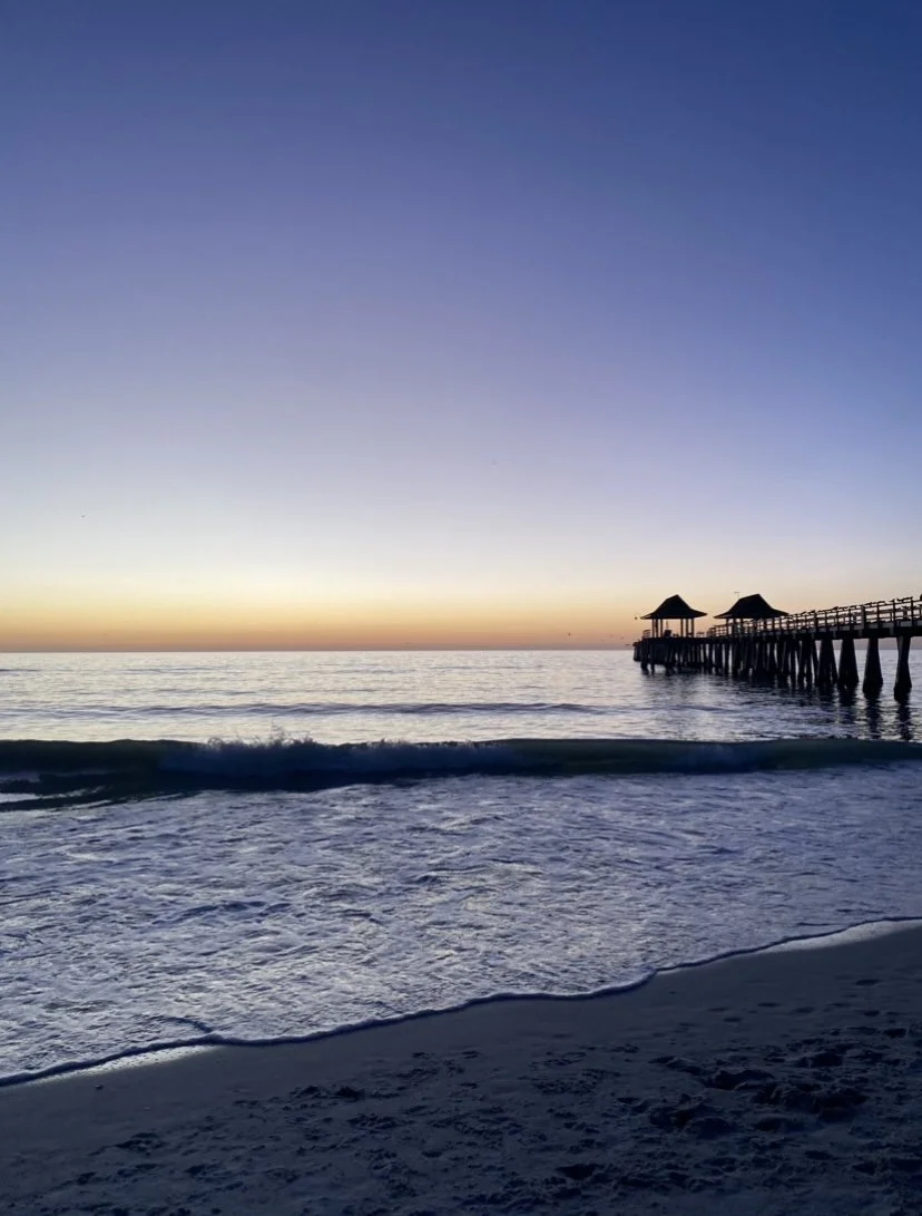 Sunset at the beach with waves and a wooden pier with small roofs extending into the water.