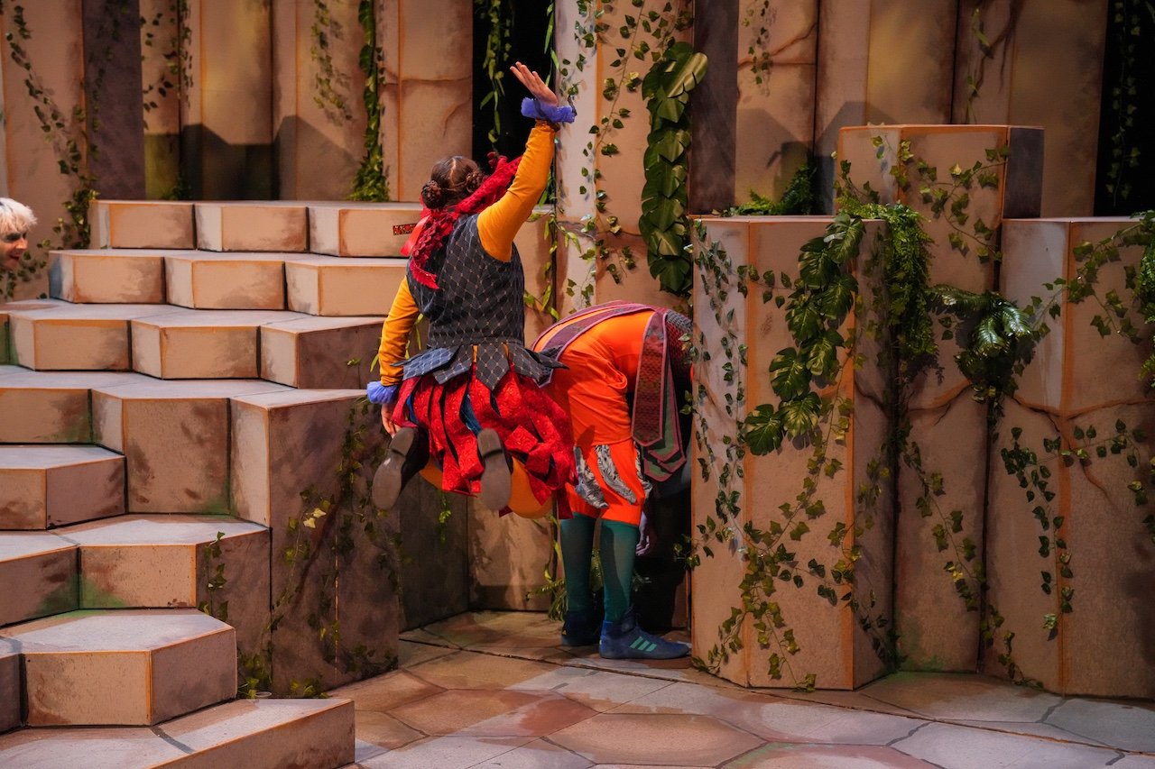 Actors dressed as Jack and the Beanstalk characters appear to be climbing or investigating a castle made of large stone blocks with ivy and greenery on the walls.