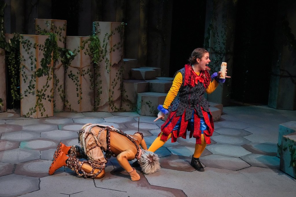 A theatrical performance with two actors on stage, one dressed as a fairy tale character on the ground and the other as a colorful storyteller standing, with set pieces resembling a forest scene in the background.