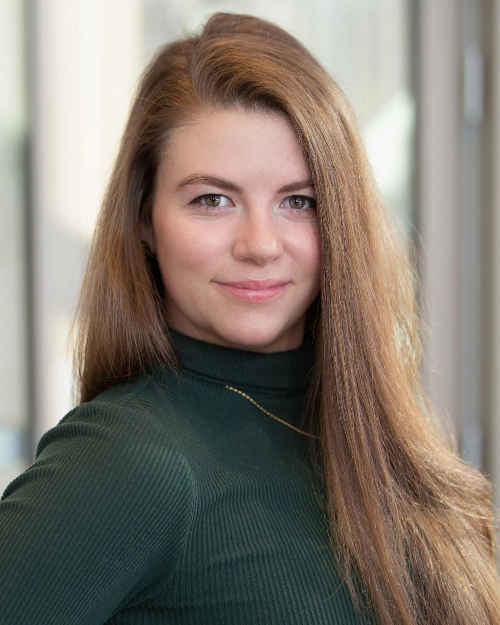 Portrait of a young woman with long auburn hair, wearing a dark green turtleneck sweater, smiling softly, indoors near a window.