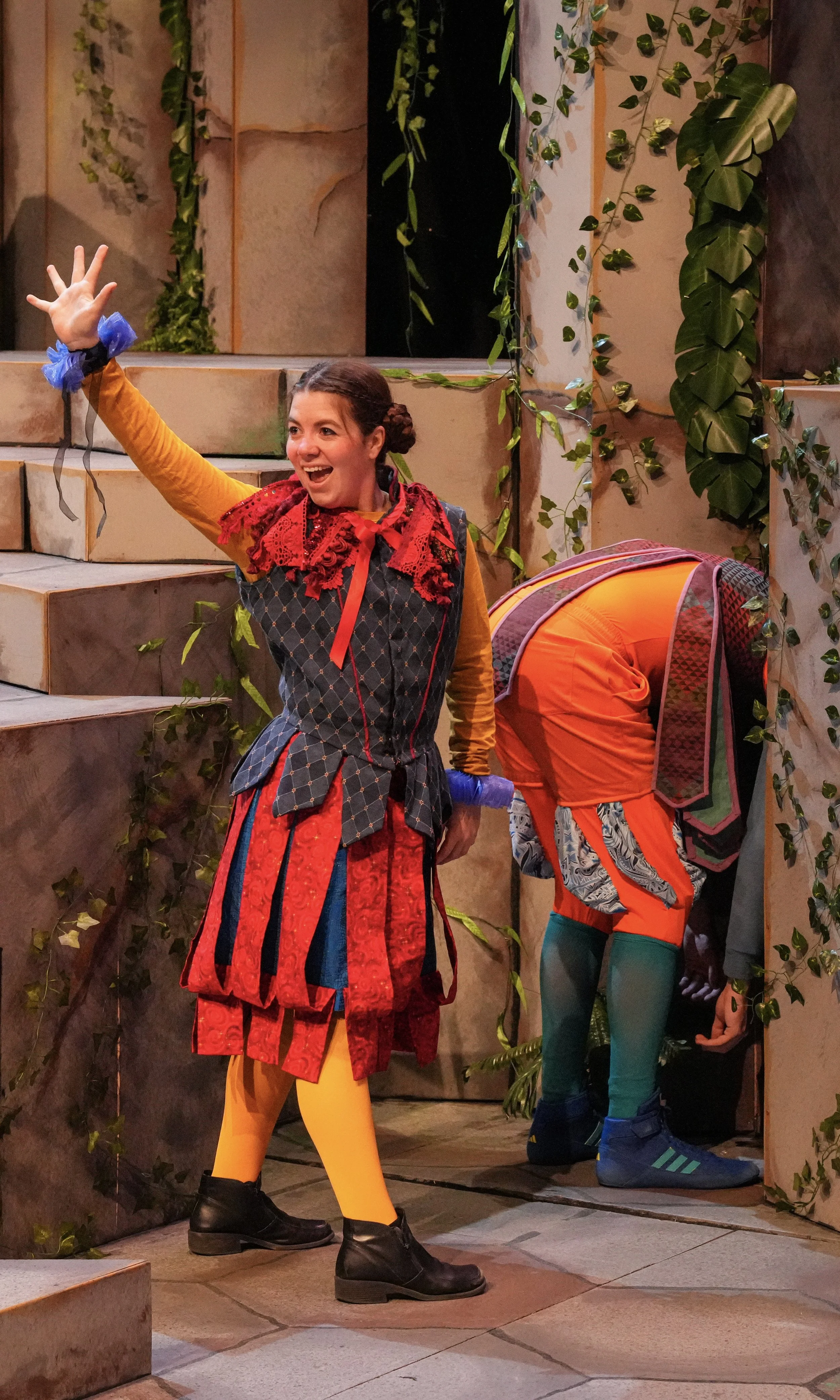 A woman in colorful, theatrical costume with a red and black patterned top, yellow tights, and black shoes, raising her hand and smiling, on stage with a woman in bright orange costume peering into a hole, surrounded by faux stone and leafy decorations.