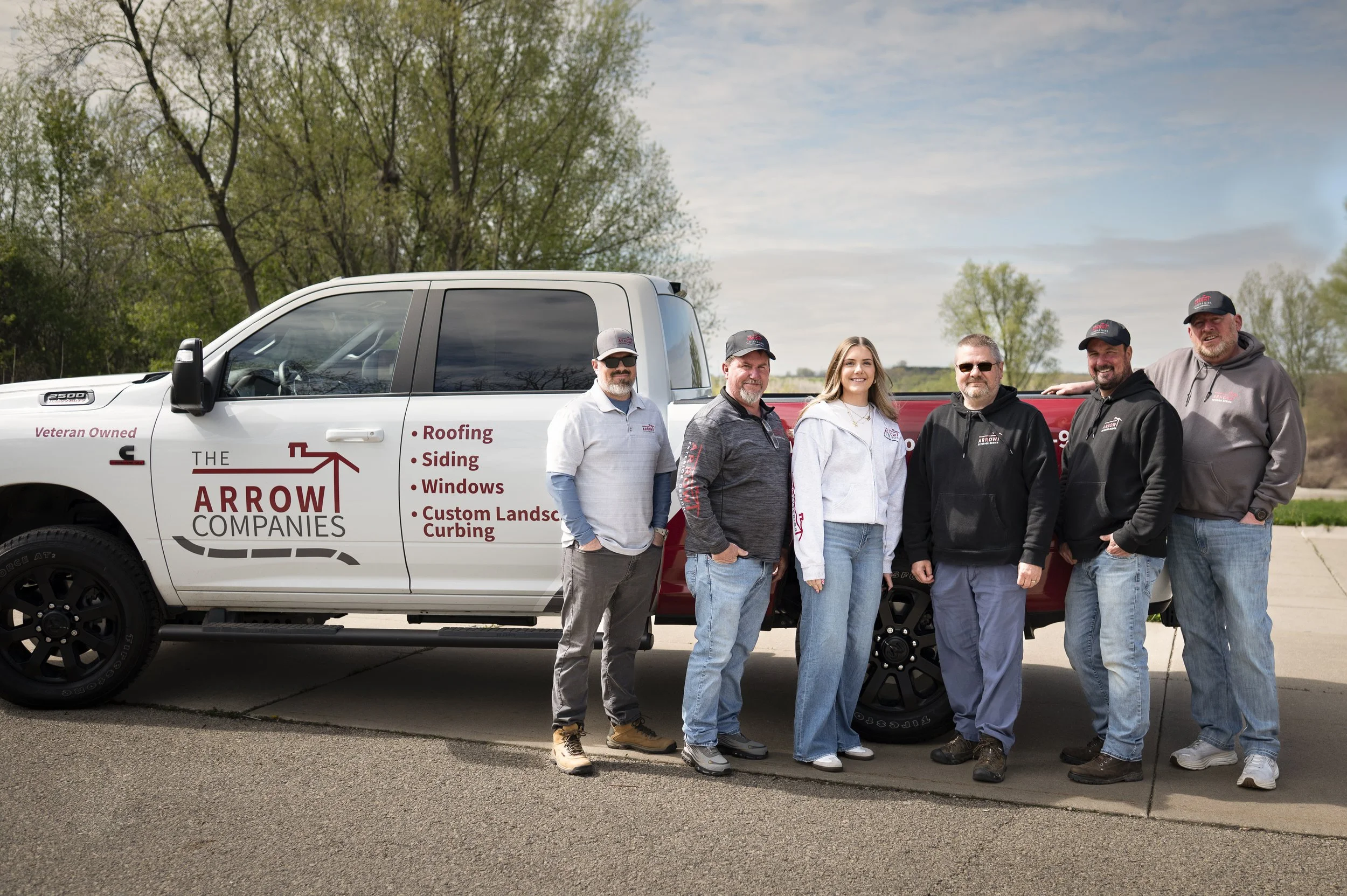 A group of six people standing beside a white pickup truck. The truck is branded with 'The Arrow Companies' logo and services such as roofing and siding. The license plate area says 'Veteran Owned.' The setting is an outdoor location with trees in the background.
