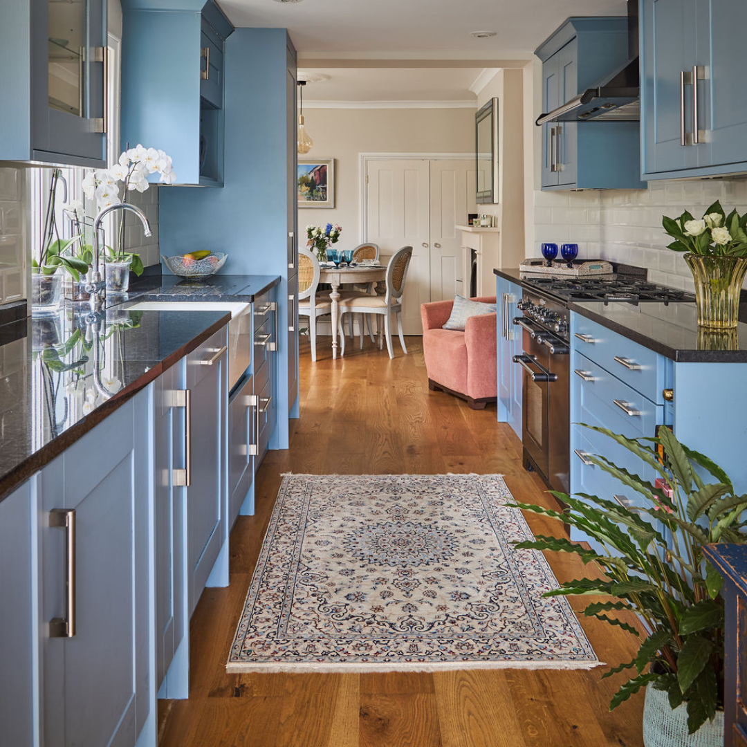 View of a cozy kitchen with blue cabinets, black countertops, a patterned rug on wooden floor, and a small dinette area with chairs in the background.