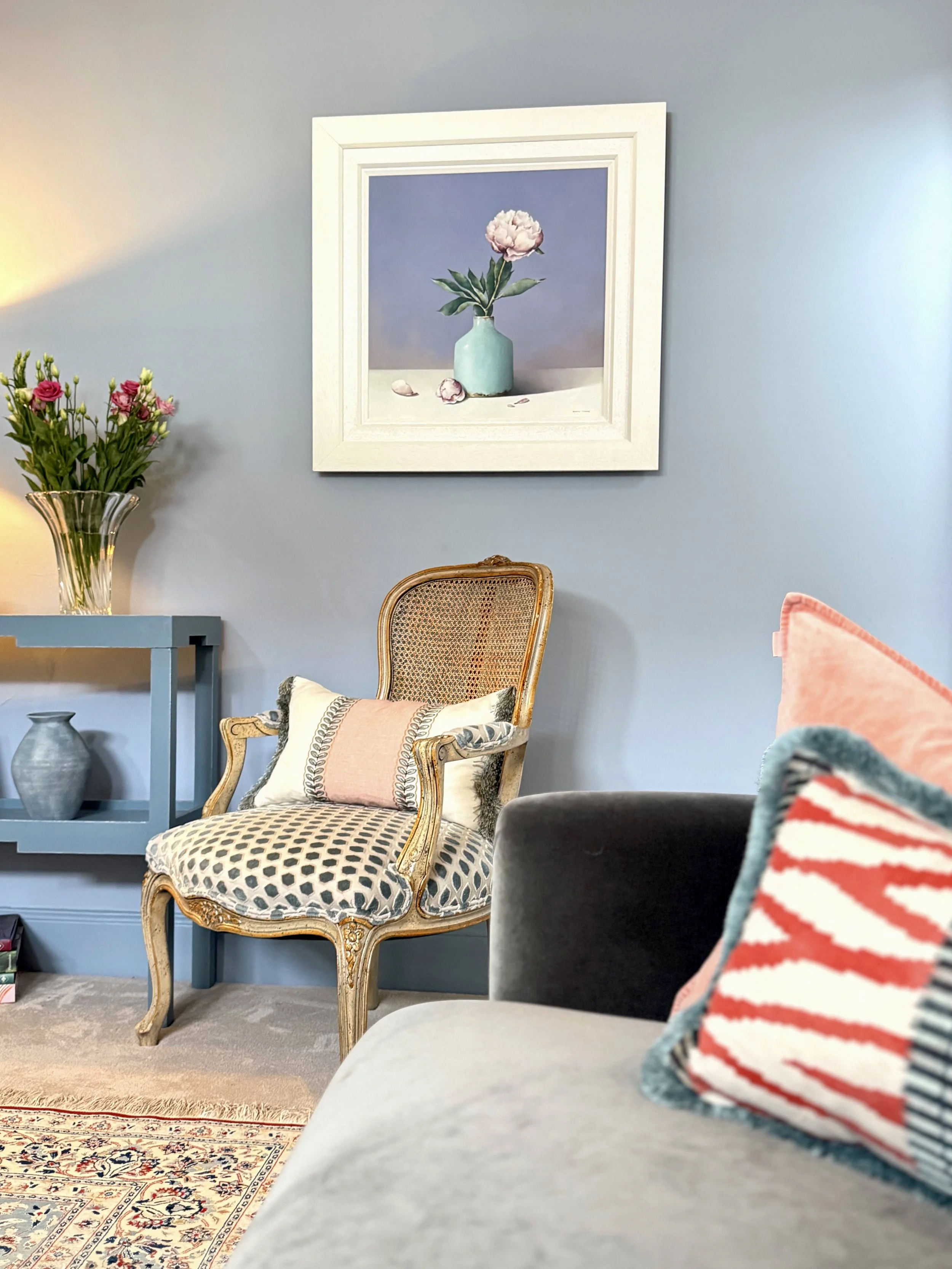 A wicker chair and above it a framed piece of art depicting a singular pink flower in a vase.