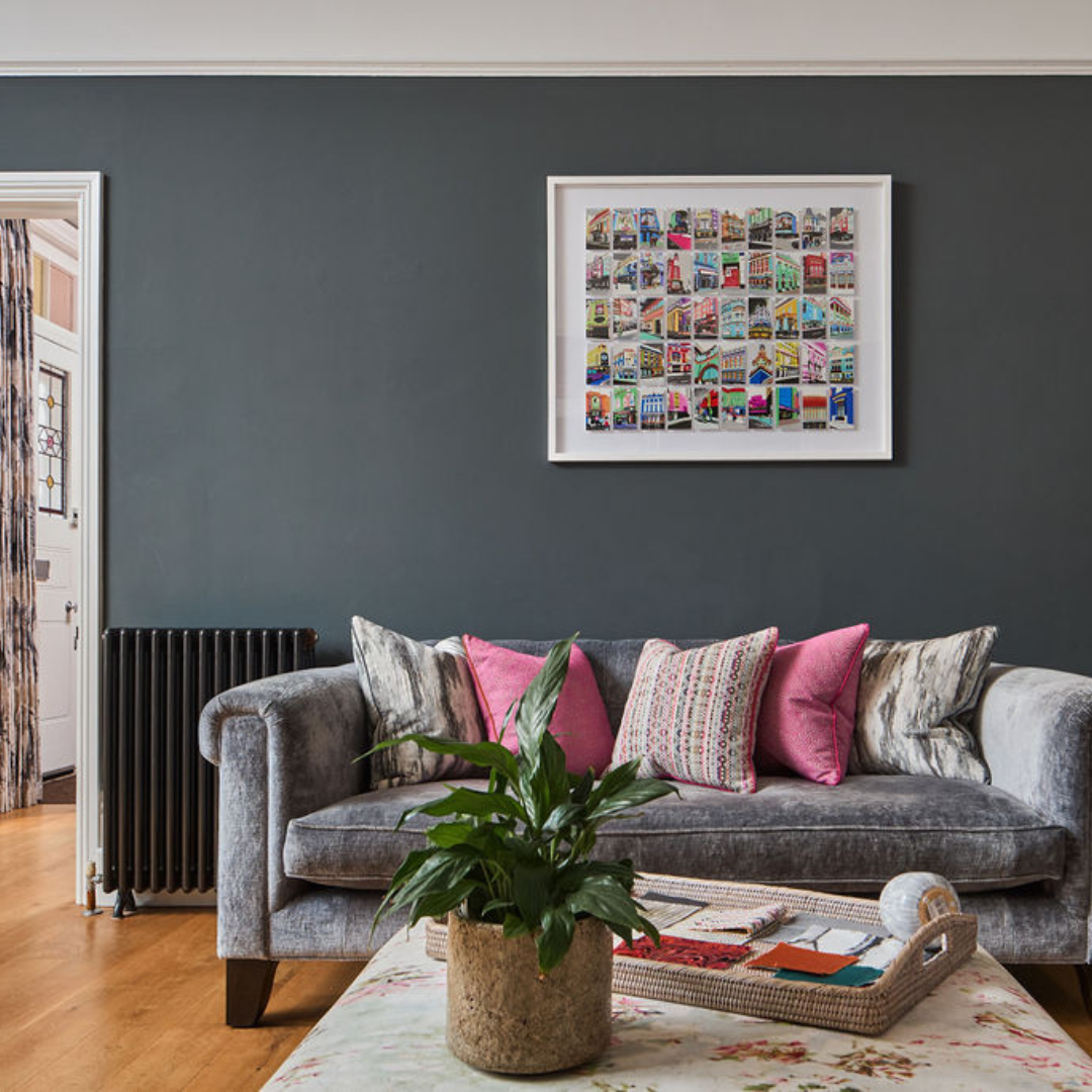 Living room with a gray sofa, pink and patterned throw pillows, a potted plant on a coffee table, framed colorful artwork on the dark blue wall, hardwood floors, and part of a door with curtain on the left.