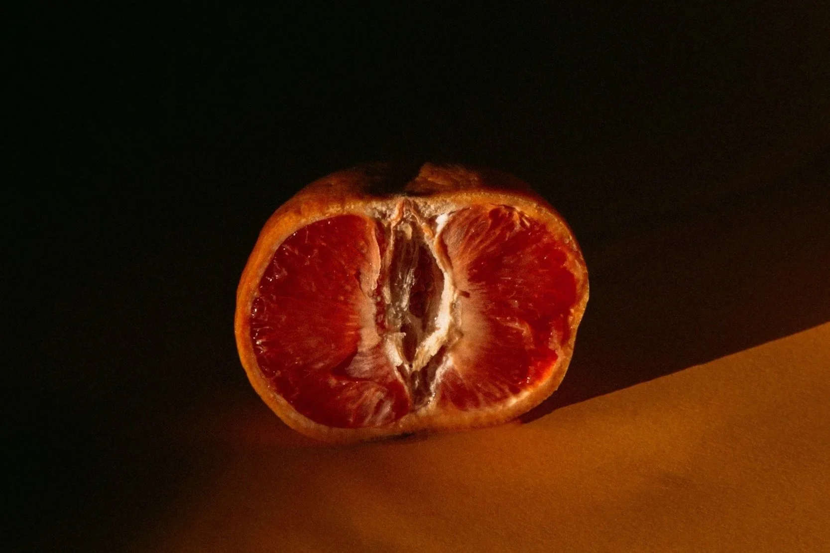 Half-cut red citrus fruit, possibly a grapefruit or blood orange, with a dark background and warm colored surface.