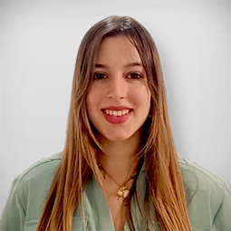 Jovem mulher sorrindo em um restaurante, com cabelo longo e ondulado, usando blusa marrom, com uma jovem ao fundo sentada à mesa.