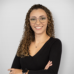 Mulher com cabelo cacheado, óculos, sorrindo, vestindo blusa preta, braços cruzados, fundo branco.
