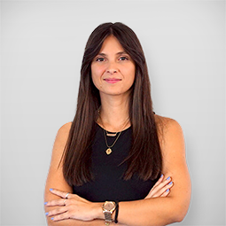 Mulher com cabelo longo e escuro, vestindo uma blusa preta, com braços cruzados, sorrindo levemente, em fundo branco.