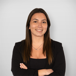 Mulher com cabelo castanho, vestindo blazer preto, sorrindo, com braços cruzados contra fundo branco.