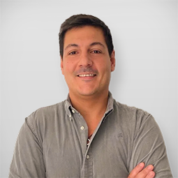 Homem sorridente com braços cruzados, de cabelo castanho escuro, usando camisa cinza, posando frente a uma parede branca.