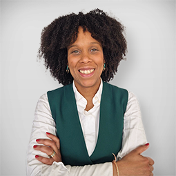 Jovem mulher sorrindo em um restaurante, com cabelo longo e ondulado, usando blusa marrom, com uma jovem ao fundo sentada à mesa.