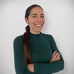 Mulher sorrindo, de cabelo longo, vestindo blusa preta, usando um colar com pingente de borboleta, tirando selfie em ambiente interno bem iluminado.