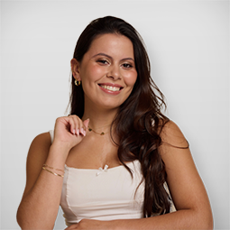 Mulher jovem sorridente com cabelos longos e castanhos, vestindo blazer azul claro e camiseta preta, contra fundo branco.