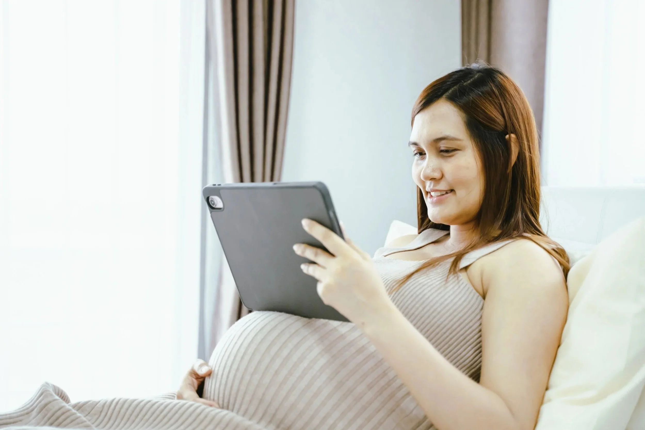 Pregnant woman using a tablet indoors, sitting on a bed near a window with curtains.
