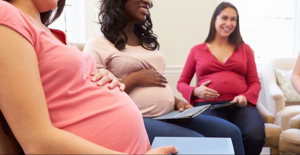 Three pregnant women sitting on a couch at a prenatal class, each holding a tablet, smiling and engaging with each other.
