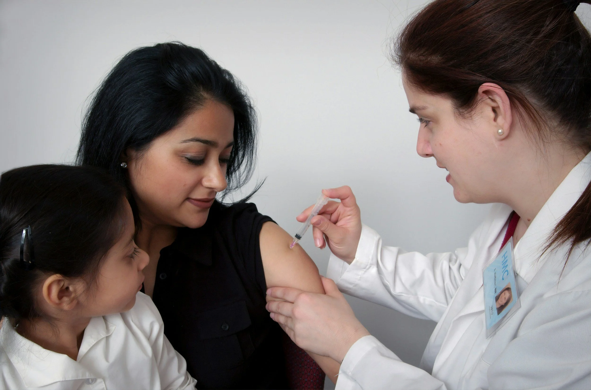A woman sits with a young girl on her lap, while a healthcare professional administers a vaccine to the woman.