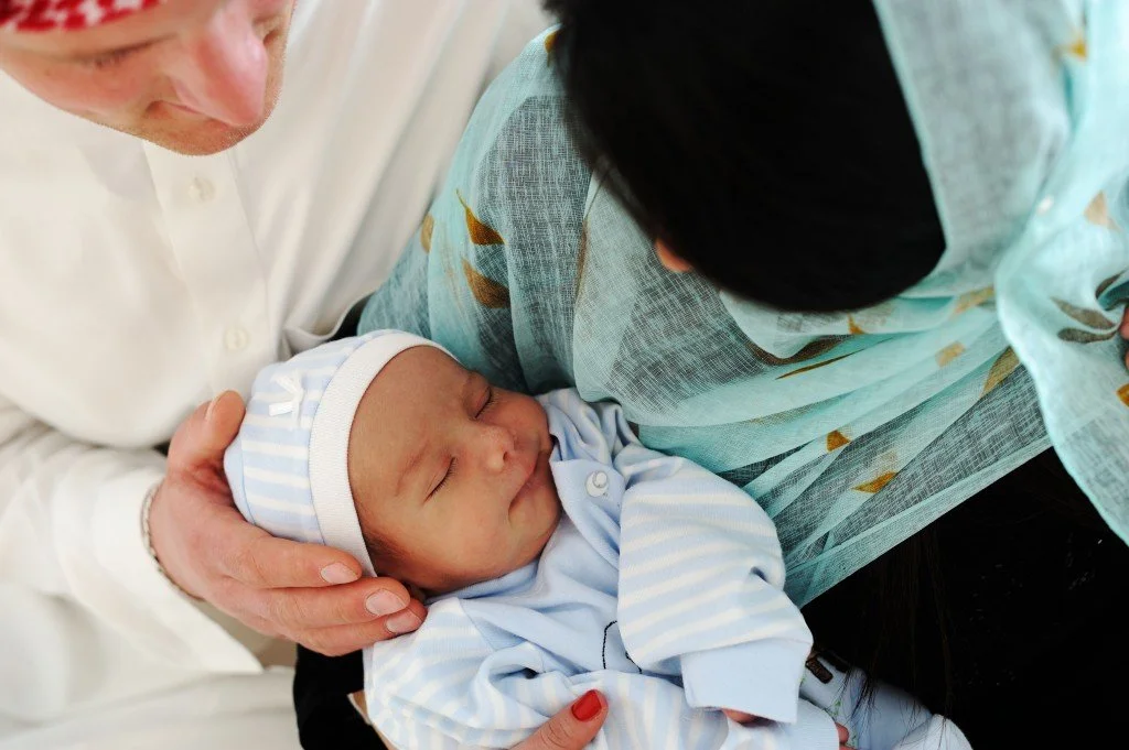 A woman wearing a hijab holding a sleeping newborn baby in her arms, while a man wearing a jubba gently touches the baby's head.