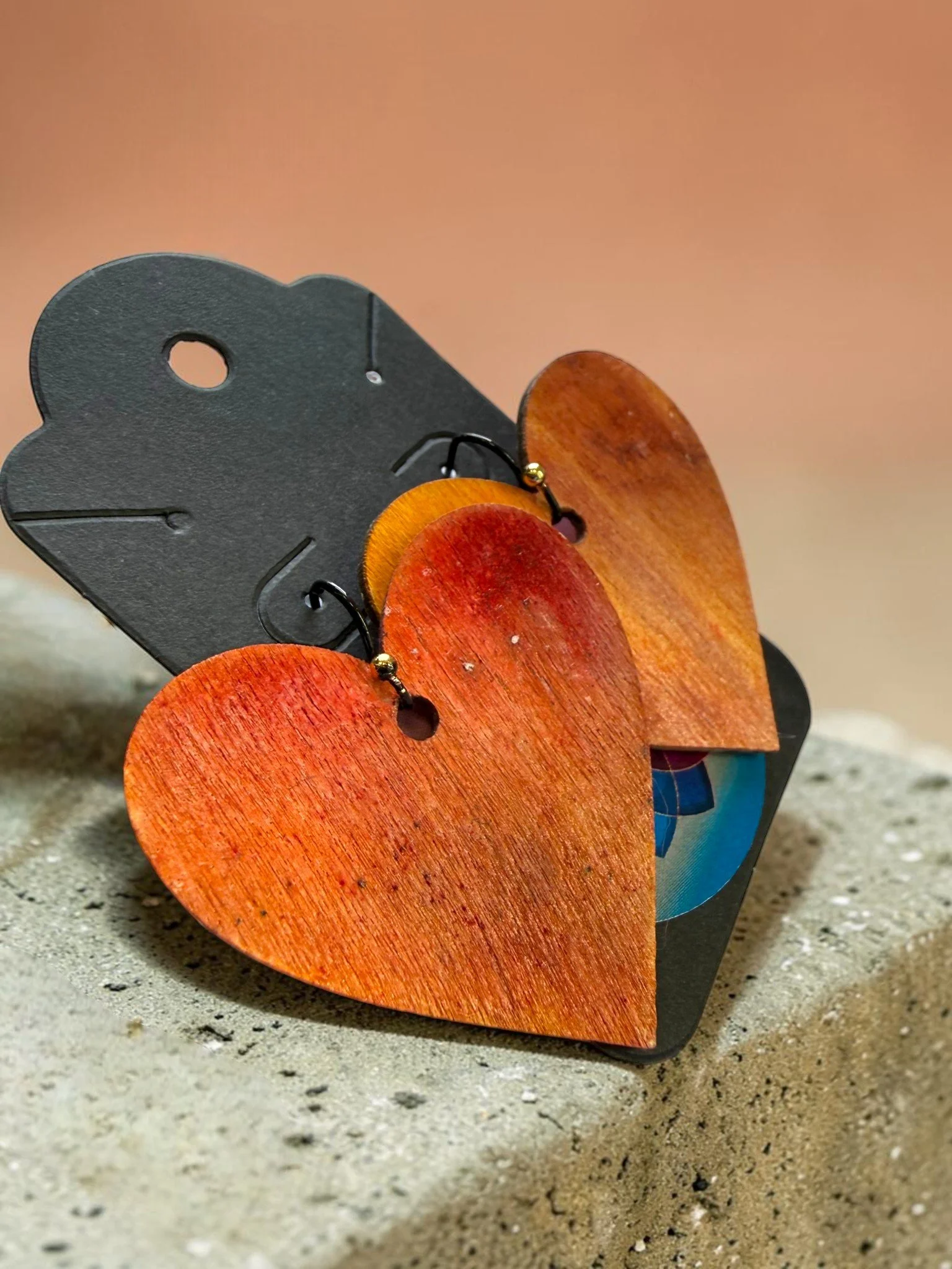 Close-up photograph of a small, decorative heart-shaped object with a red wooden surface and black hooks, placed on a textured, sandy surface with a neutral background.
