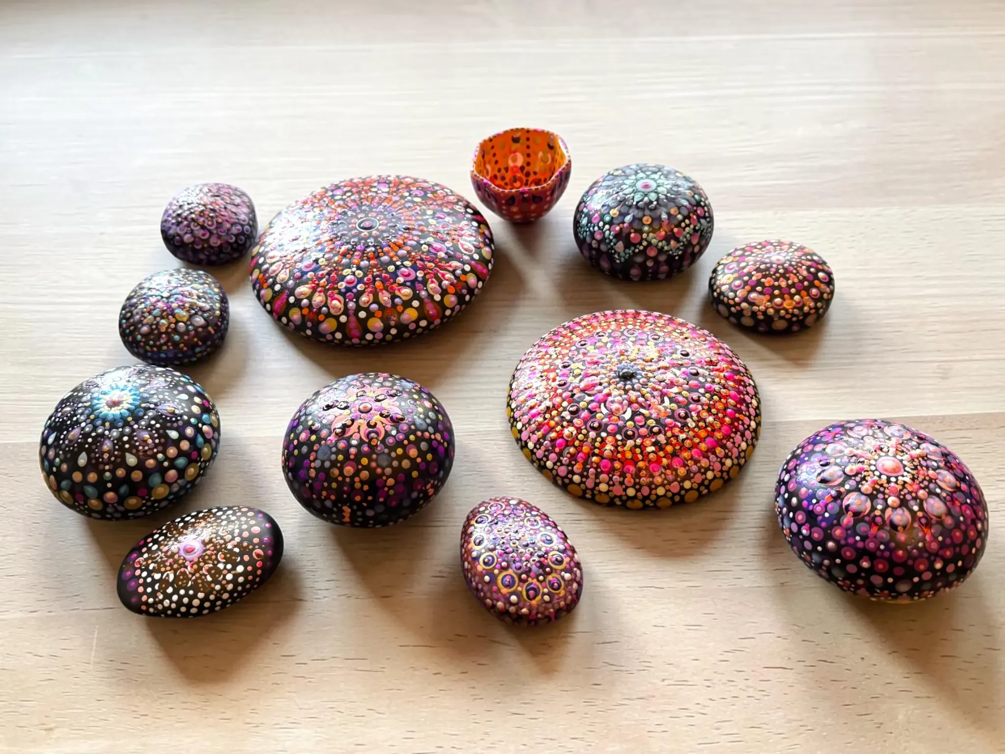A display of colorful mandala stones made by Color Cult. The stones feature colorful dot mandala designs in many colors. The stones are displayed on a wooden table.
