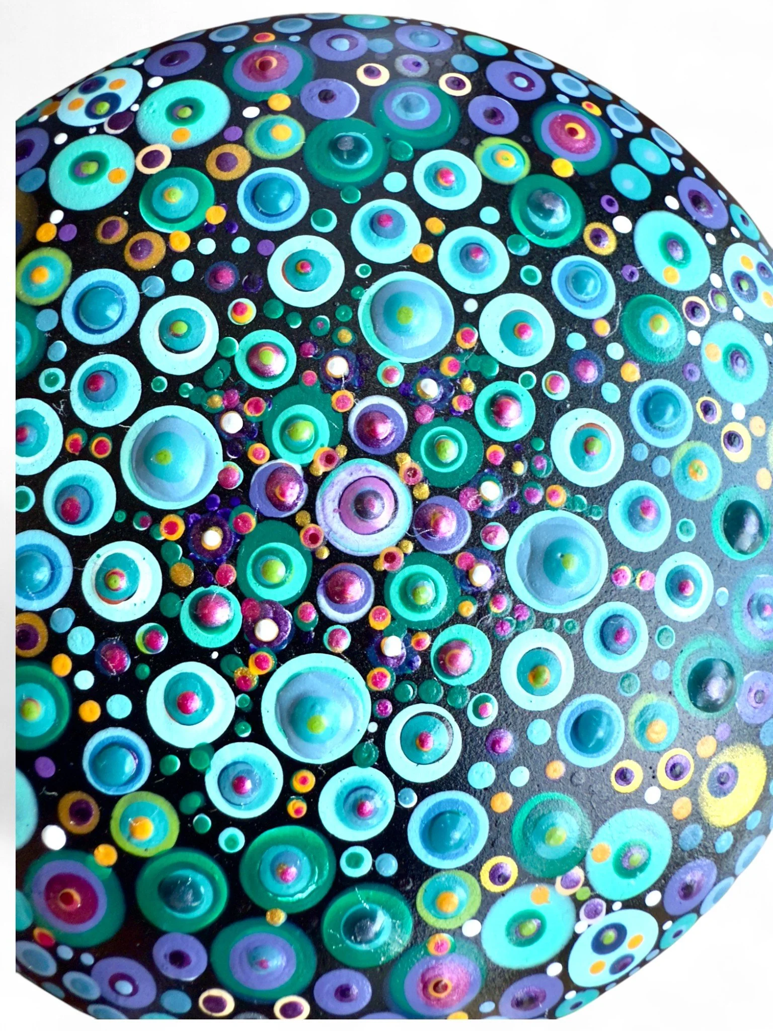Close-up of a painted rock with colorful concentric circles and dots in blue, purple, pink, yellow, and green on a black background.