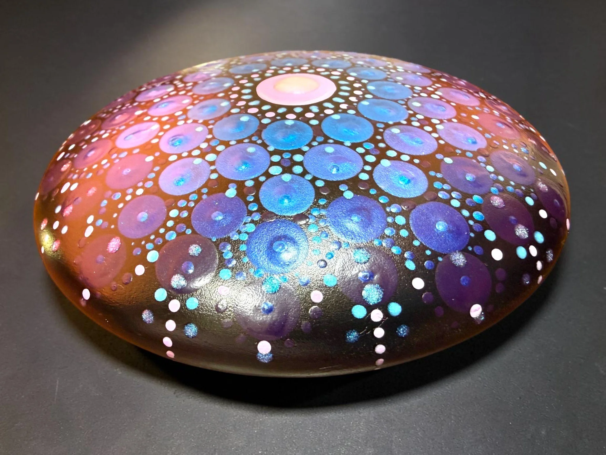 A colorful glass or stone object with a pattern of pink, blue, and purple circles and dots on its surface, resting on a dark surface.