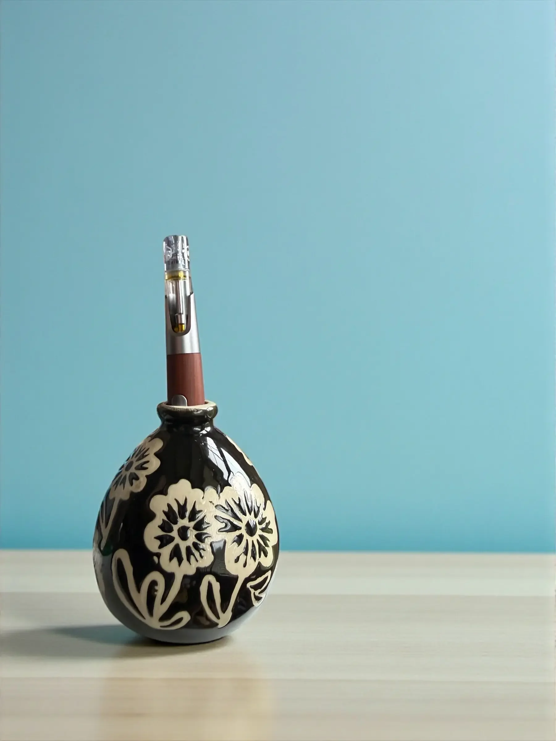 A decorative ceramic vase with a black and white floral pattern, with a wooden vape pen held inside the vase, and placed on a wooden surface against a light blue background.