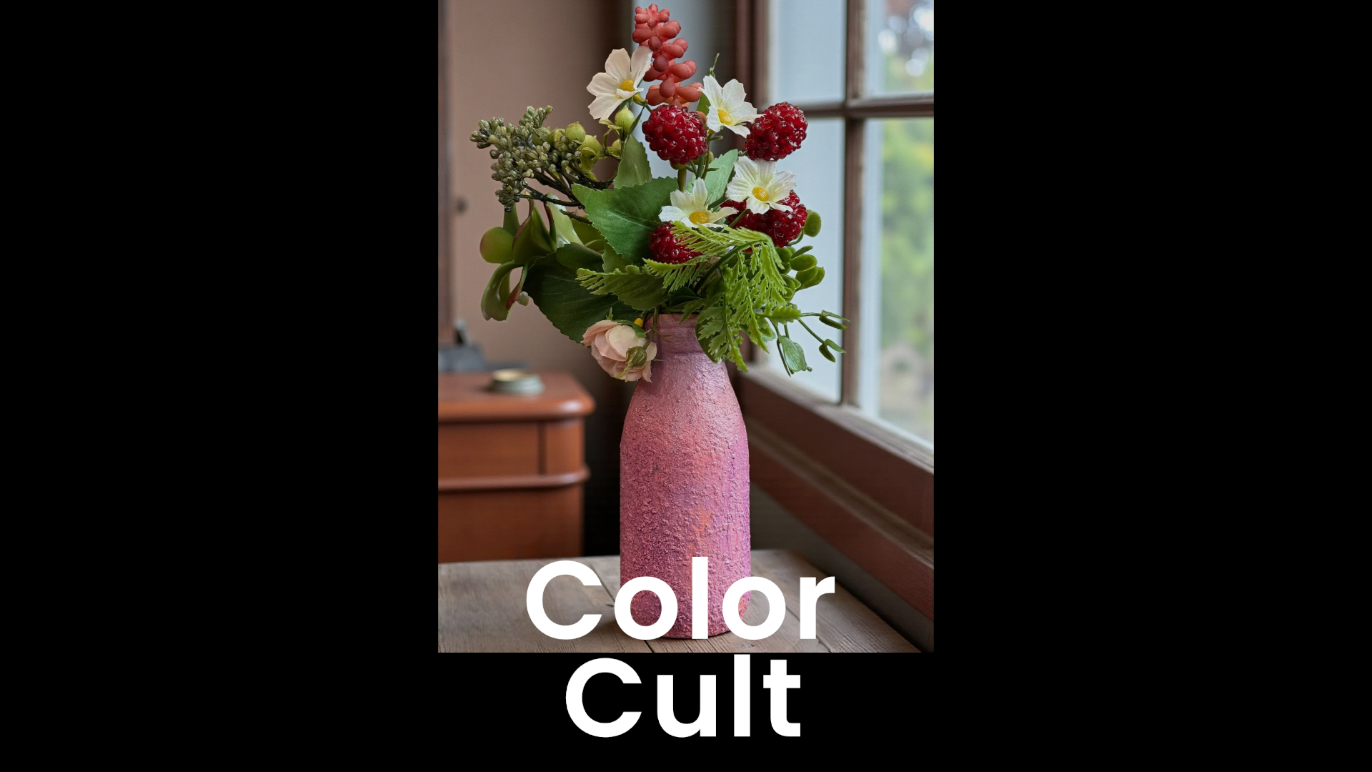 A pink textured ceramic vase with a variety of colorful artificial flowers and green leaves, placed on a wooden surface near a window with a view of the outdoors.