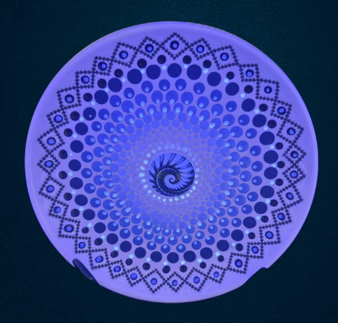 A circular, spiral-shaped decorative plate with a glass cabochon in the center, featuring a blue and black pattern with dotted and geometric designs, illuminated with a purple glow from a blacklight, to emphasize the glow in the dark features.