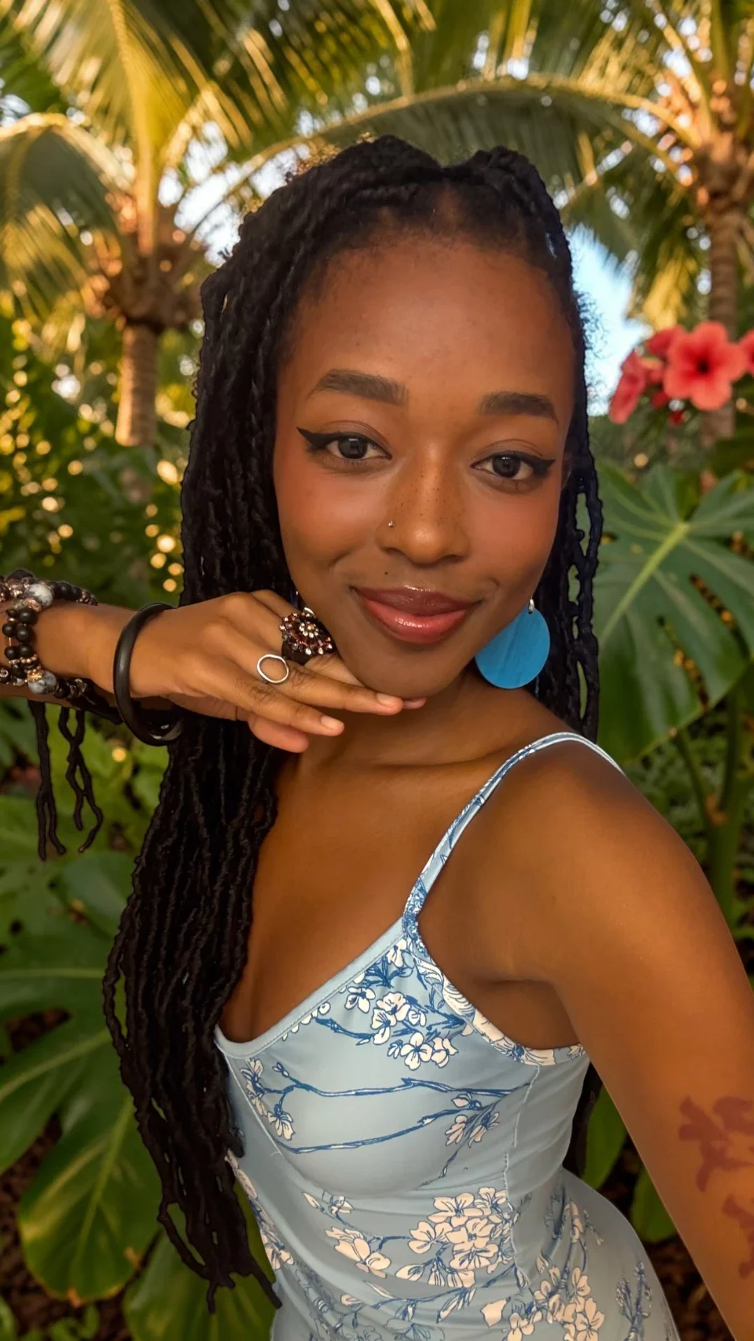 Young woman taking a selfie outdoors among tropical plants, wearing a floral dress and blue earrings, with long dreadlocks, makeup, and a nose piercing.