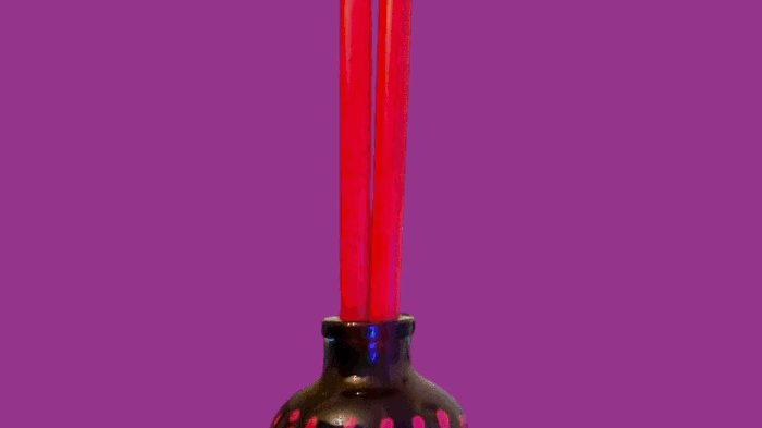 A black vase with pink dots holding red incense sticks against a purple background.