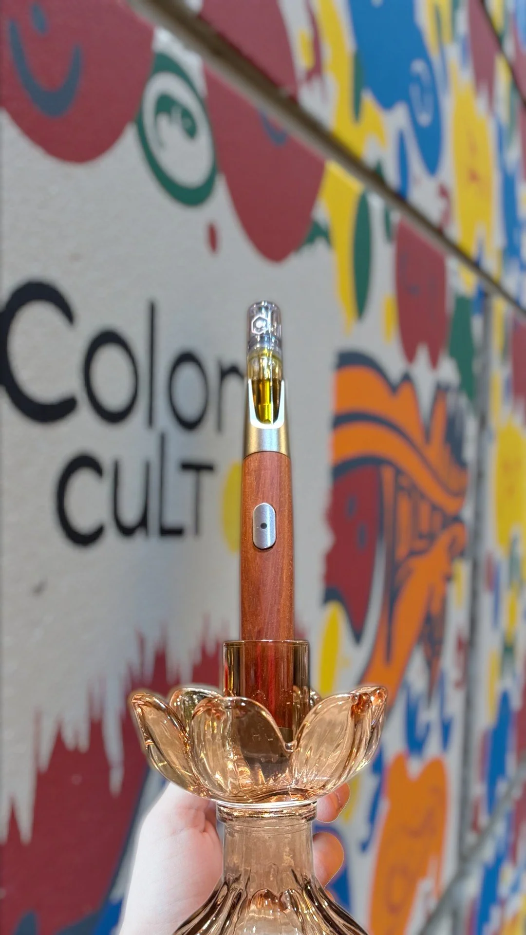 A hand holding a glass vape penholder with a wooden vaporizer or e-cigarette inserted into it, with a colorful graffiti-style wall with abstract designs and partially visible text in the background.