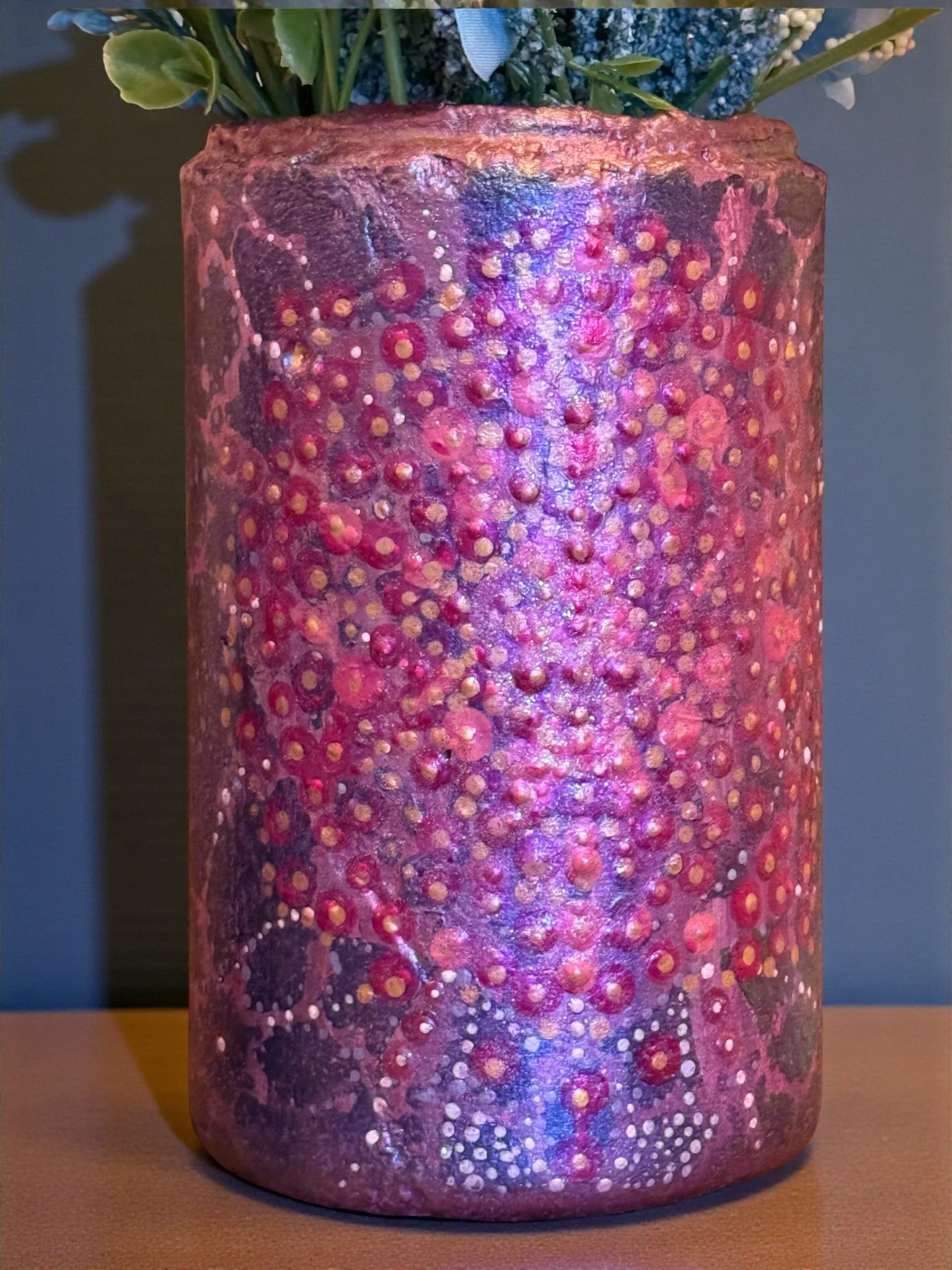 A tall, cylindrical vase painted with an abstract pattern of pink, purple, and red dots and splashes, holding blue and white flowers.