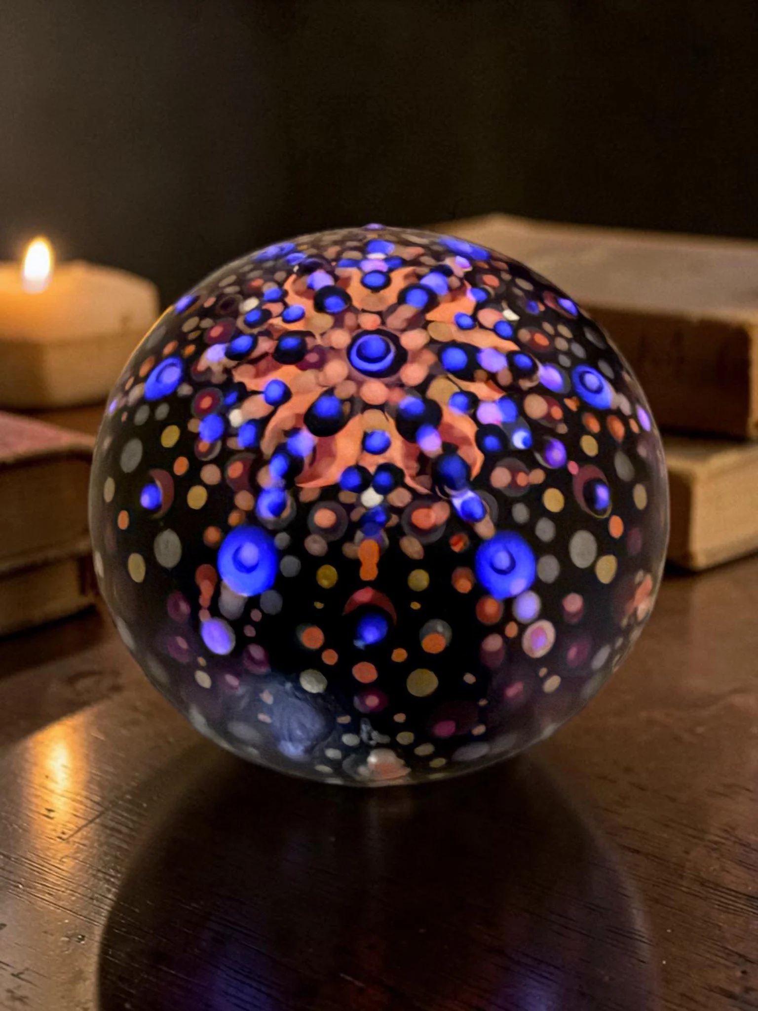 Decorative mandala stone with colorful dotted patterns, featuring some glow in the dark highlights, placed on a wooden surface with candles in the background.