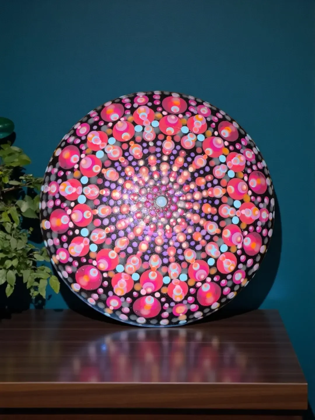 Colorful mandala stone made by Color Cult with pink, purple, orange, and blue dots on a black background, resembling a kaleidoscope mandala design, placed on a wooden surface with a green plant on the left.