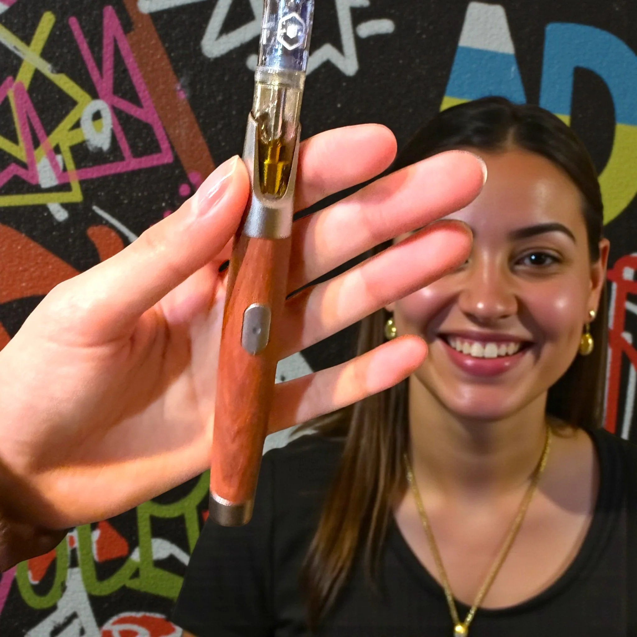 Woman with long brown hair smiling and wearing gold earrings and necklace taking a selfie while holding a vape pen in front of a colorful graffiti wall.