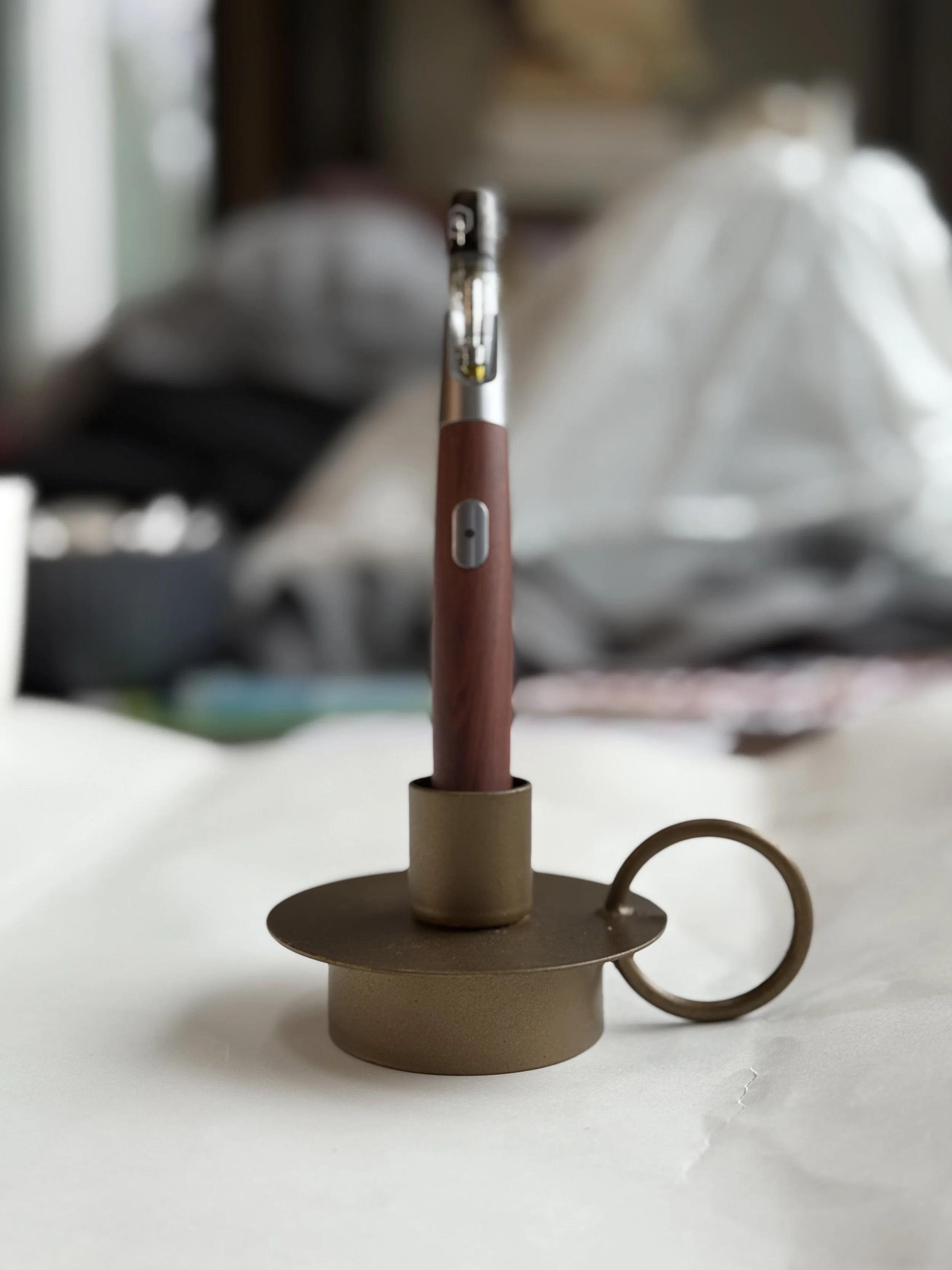 A tablet or e-reader with a wooden case, placed on a decorative stand with a circular top and ring handle, on a white surface, with blurry background of household items.