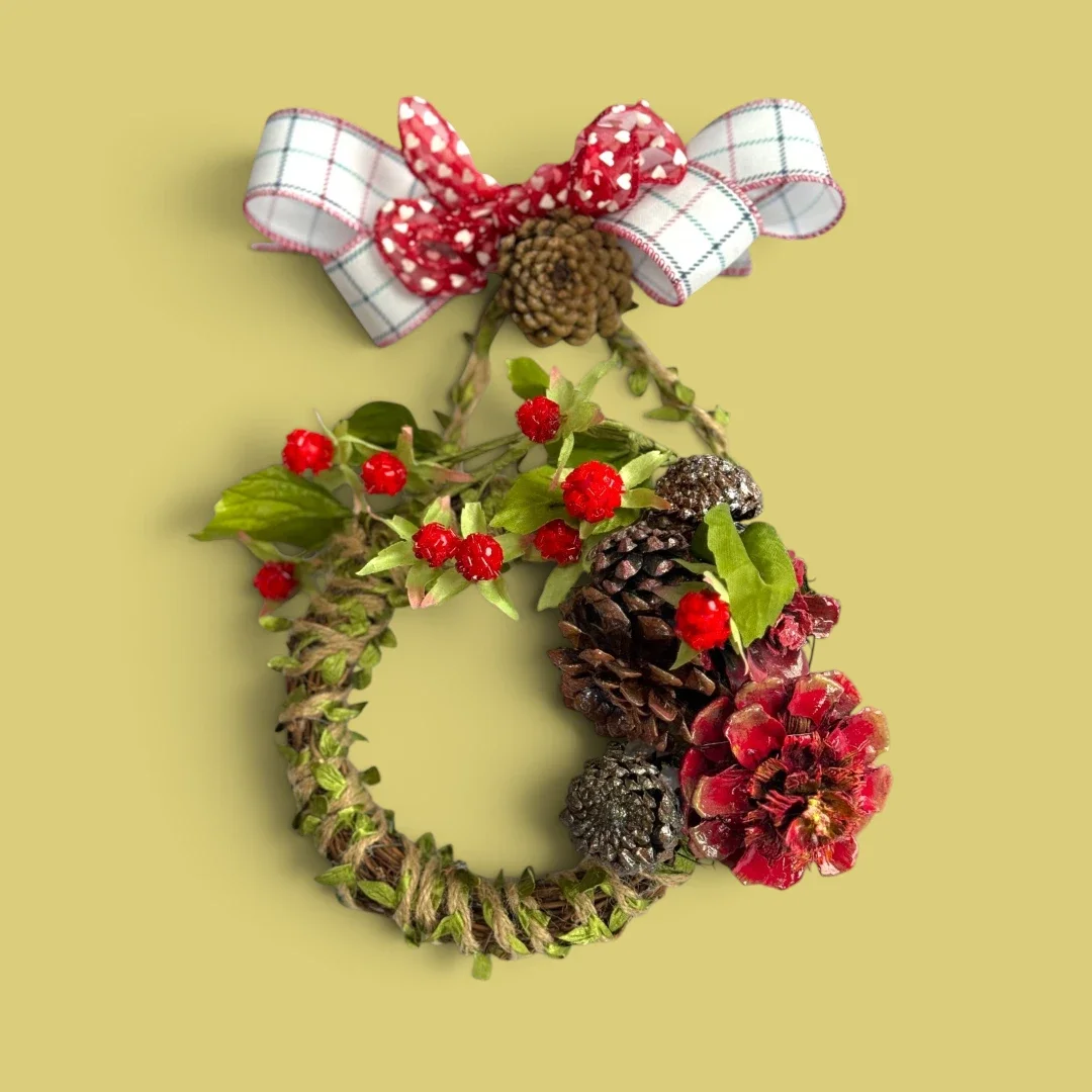 A small wreath made of hand-sculpted pinecones made to look like succulents and a flower. There are two bows at the top of the wreath and some faux greenery and berries around the wreath.