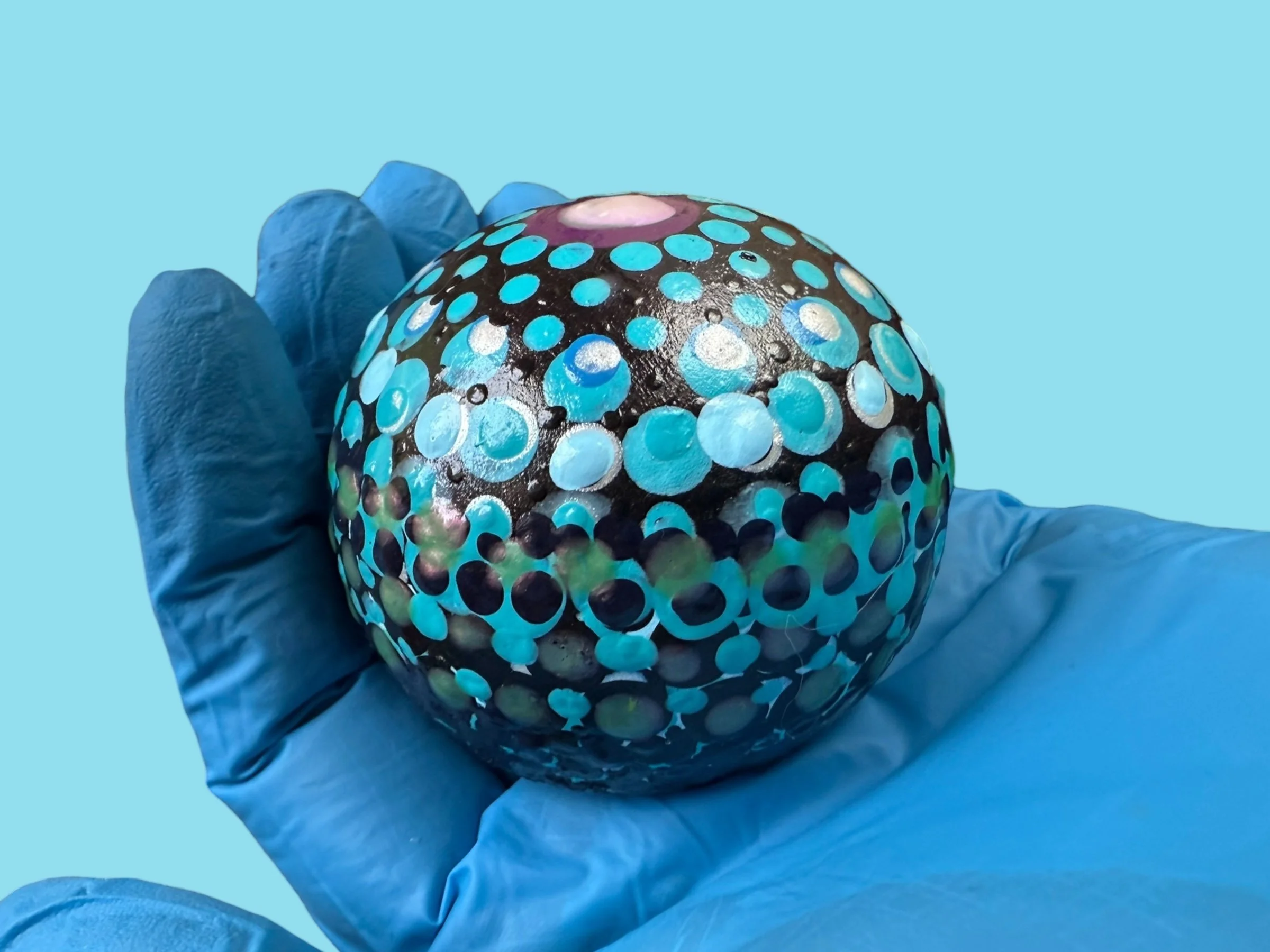 A gloved hand holding a spherical object painted with blue, black, and purple dots.