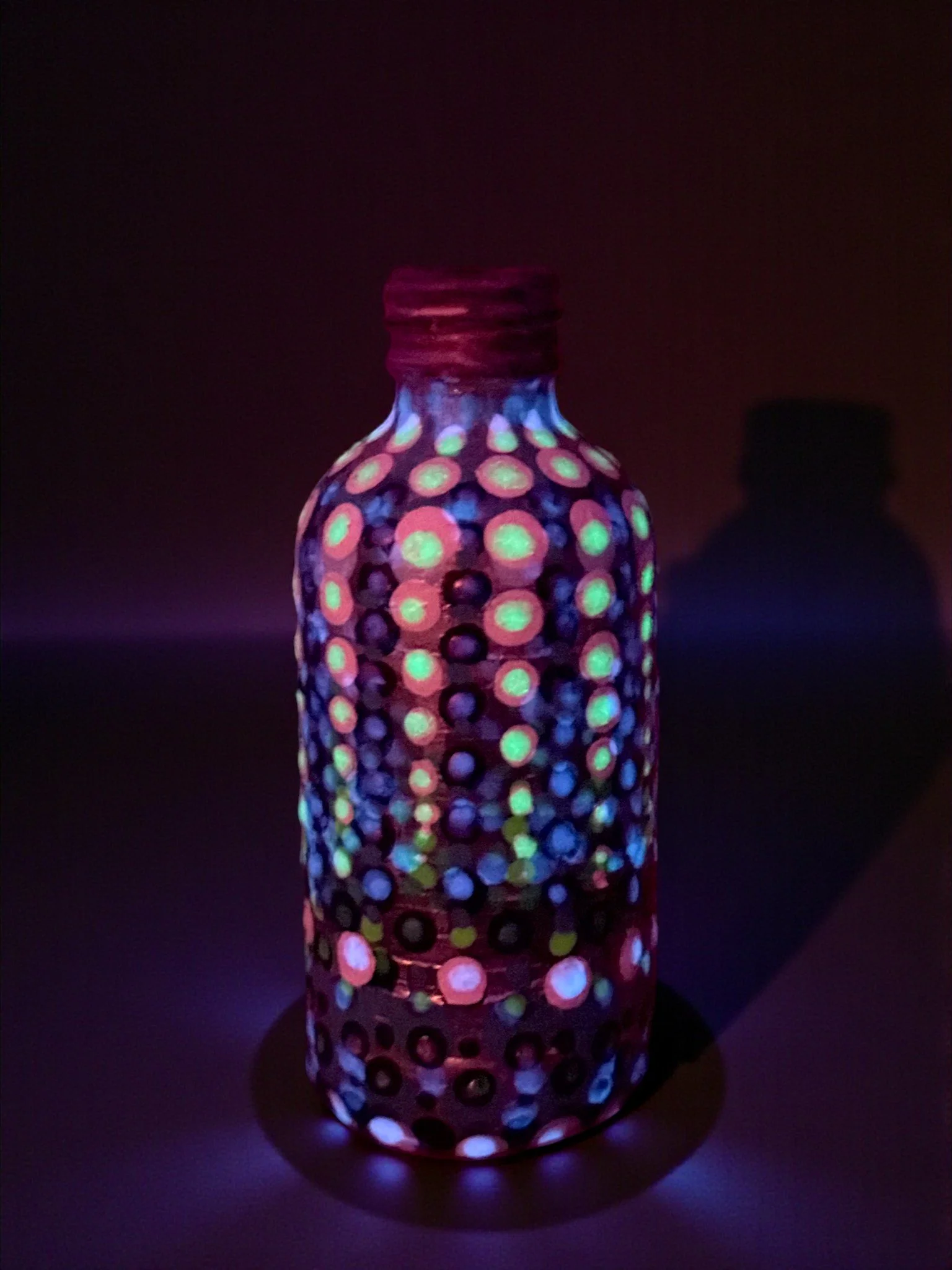 Glass bottle with dotted patterns glowing under UV light, casting colorful spots on the surface underneath.