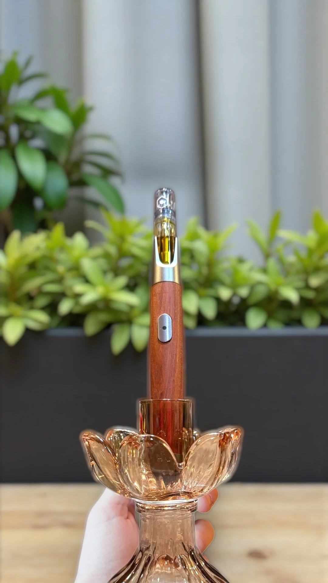 A vaporizer pen with a wooden body inserted into a decorative glass holder, with a blurred green plant and curtains in the background.