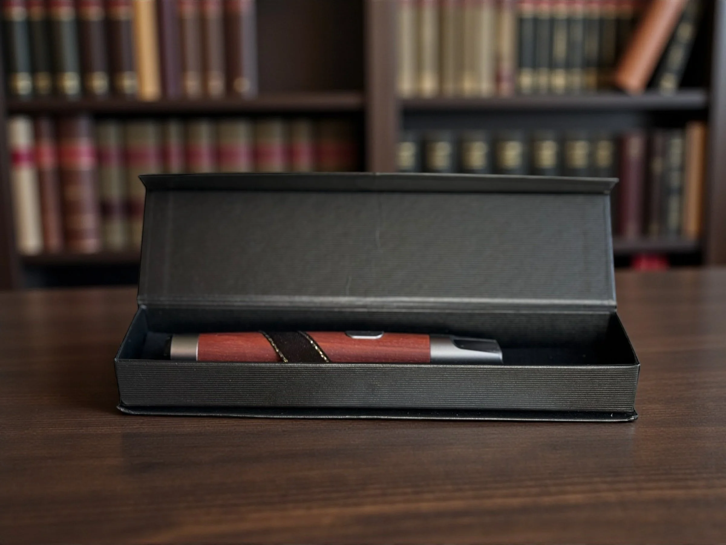 A black box with a pen inside, resting on a wooden surface. The background features a blurred bookshelf filled with books.