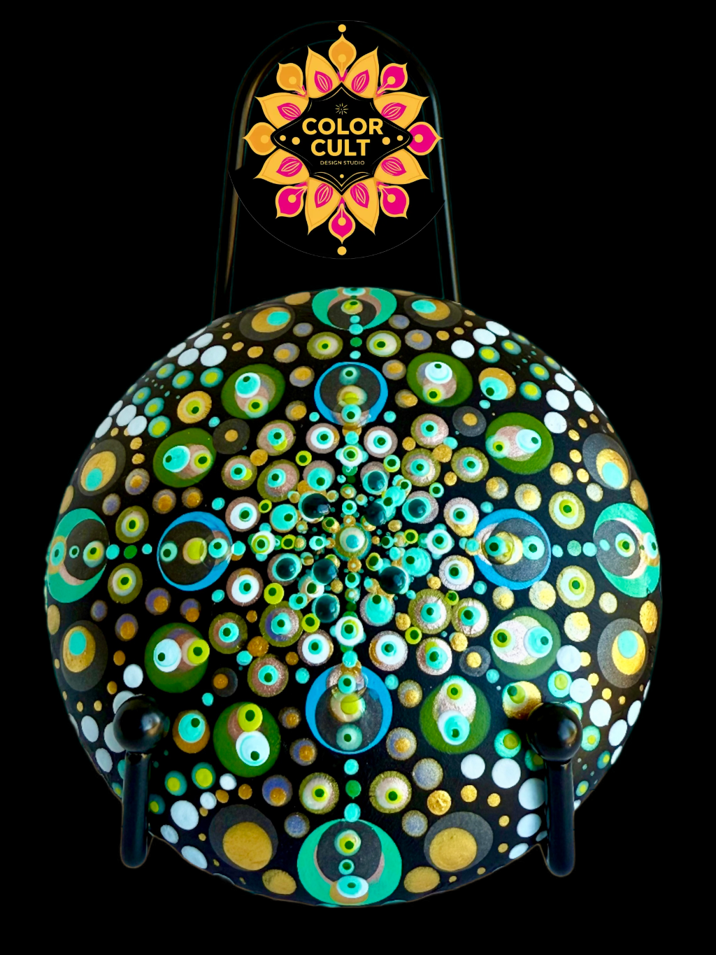 A colorfully hand-painted cement mandala stone with a pattern of multicolored concentric circles on a black background, with a sign reading 'Color Cult Design Studio' above it.