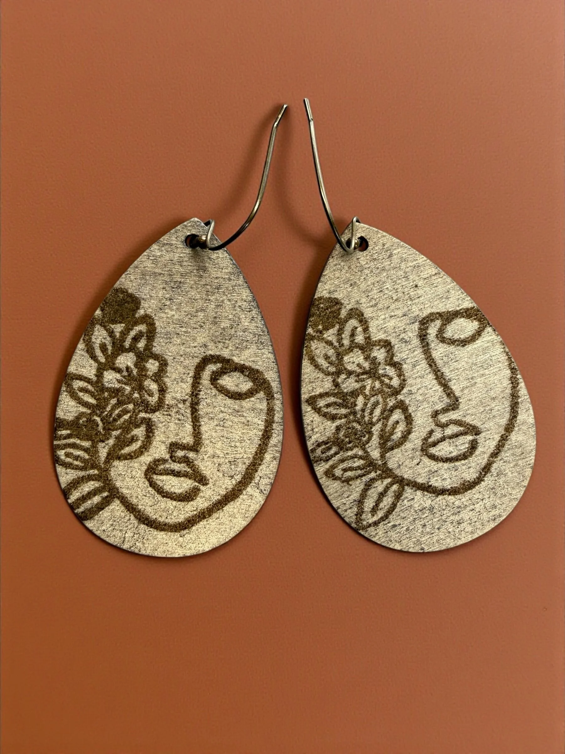 Gold-colored teardrop-shaped earrings with an embossed line drawing of a woman's face and floral details, hanging on hooks.