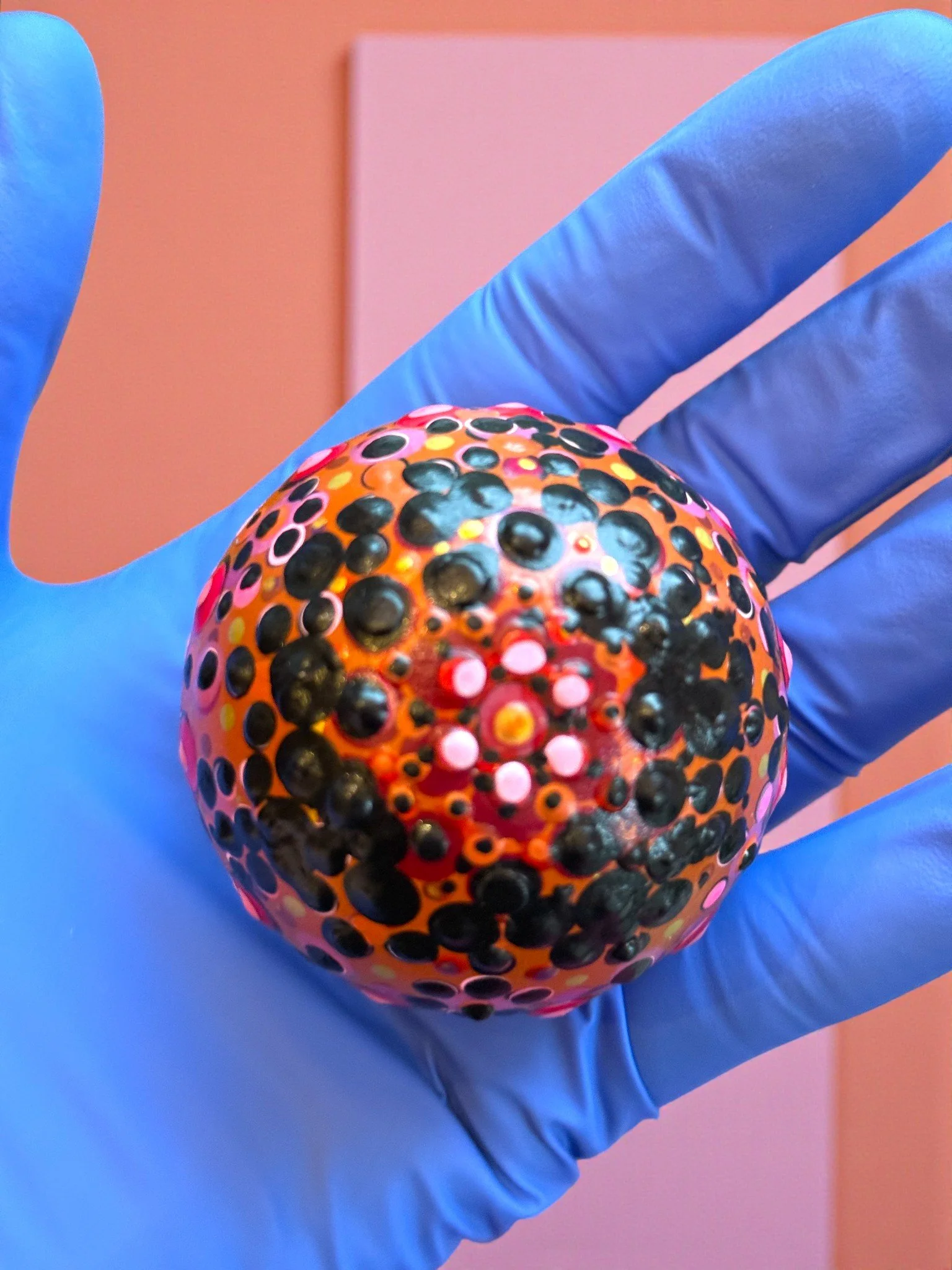 Person wearing a blue glove holding a spherical object with colorful dots and patterns, set against a pastel-colored background.