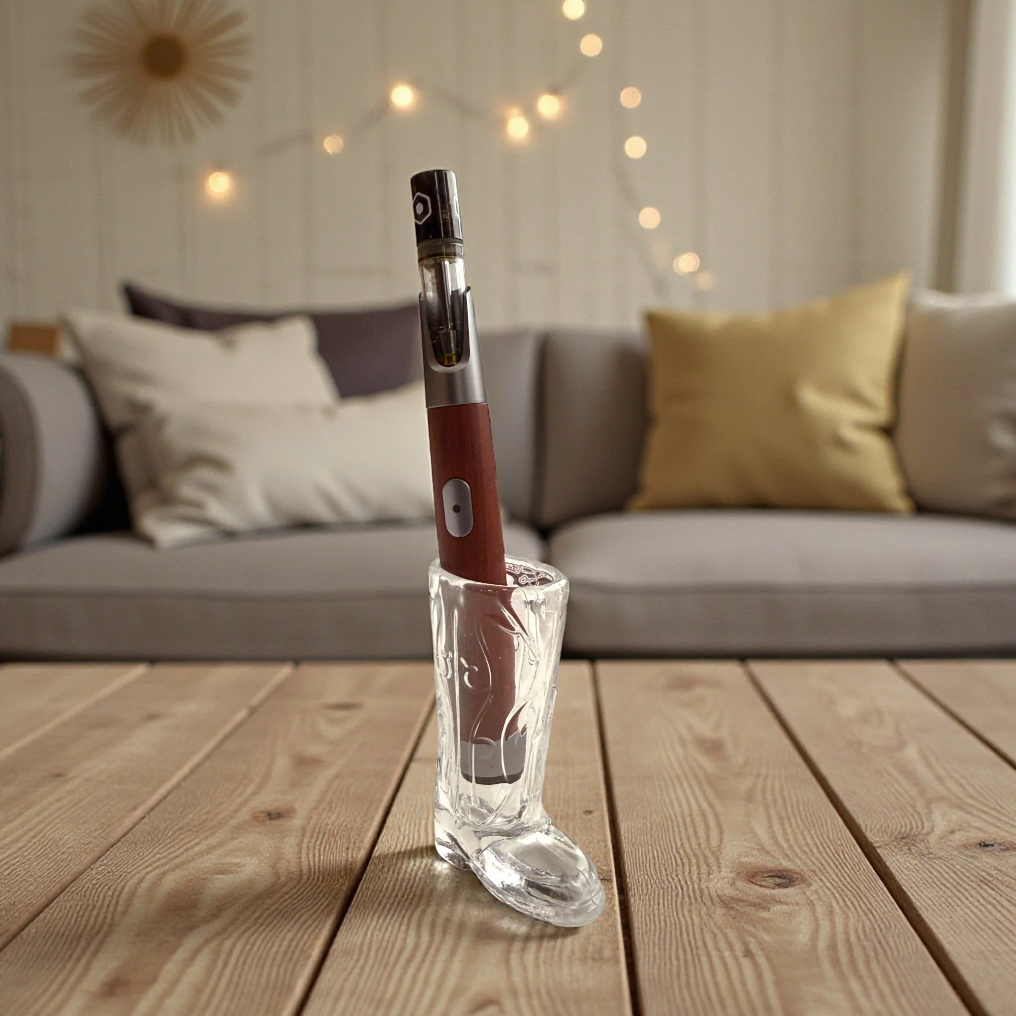 A vape pen in a glass shoe-shaped holder on a wooden table with a couch and decorative pillows in the background.
