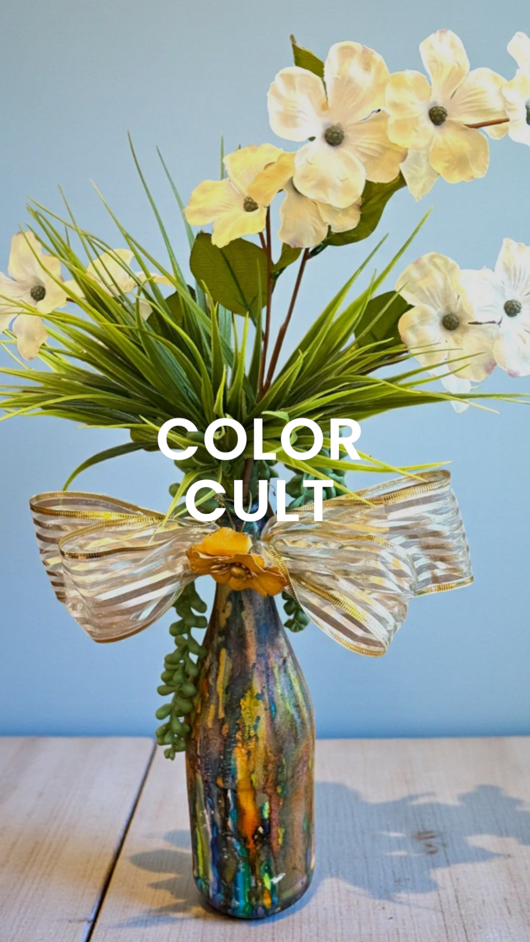 A colorful glass vase with abstract patterns holding a bouquet of white flowers, green leaves, and greenery, tied with a striped ribbon. Text overlay says 'COLOR CULT'.