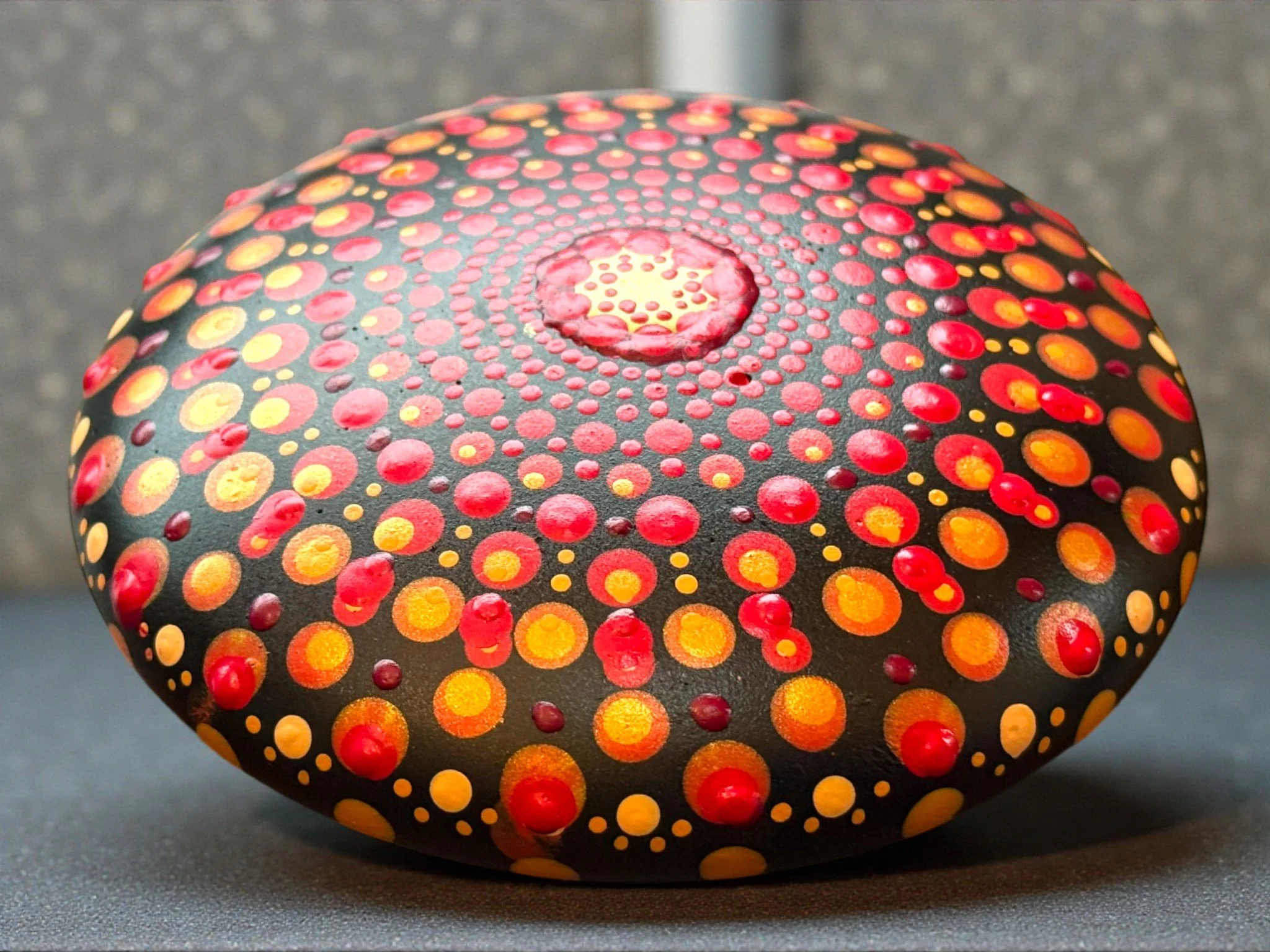 Colorful painted rock with concentric circles of red, orange, and yellow dots on a black background.