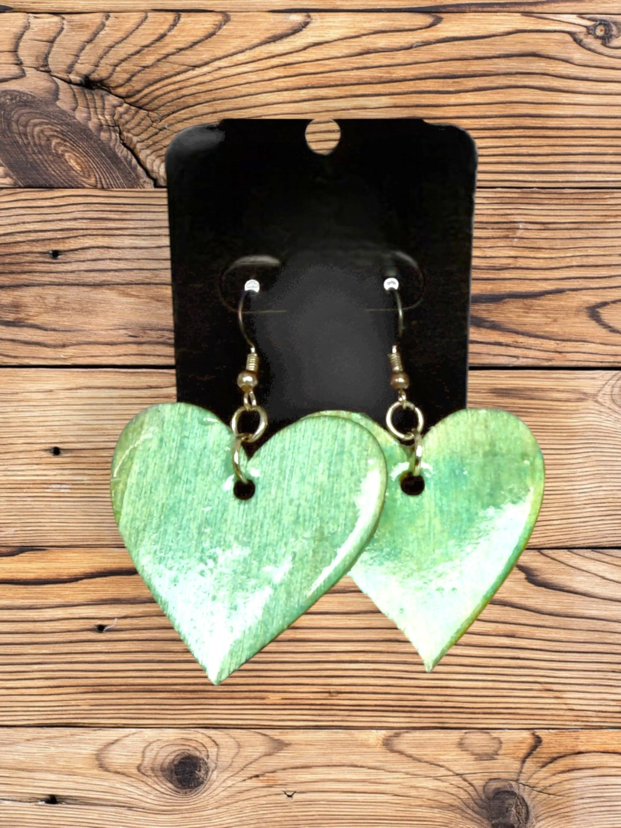 A pair of large green heart-shaped earrings with gold hooks sitting on a black stand, placed on a wooden surface.